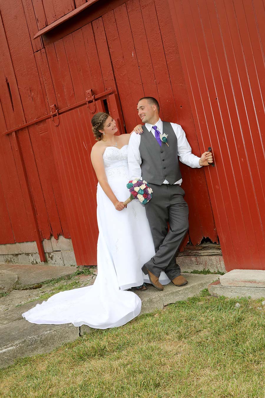 the bride is leaning on her groom as he is leaning on the red barn door on their Bowling Green Ohio farm