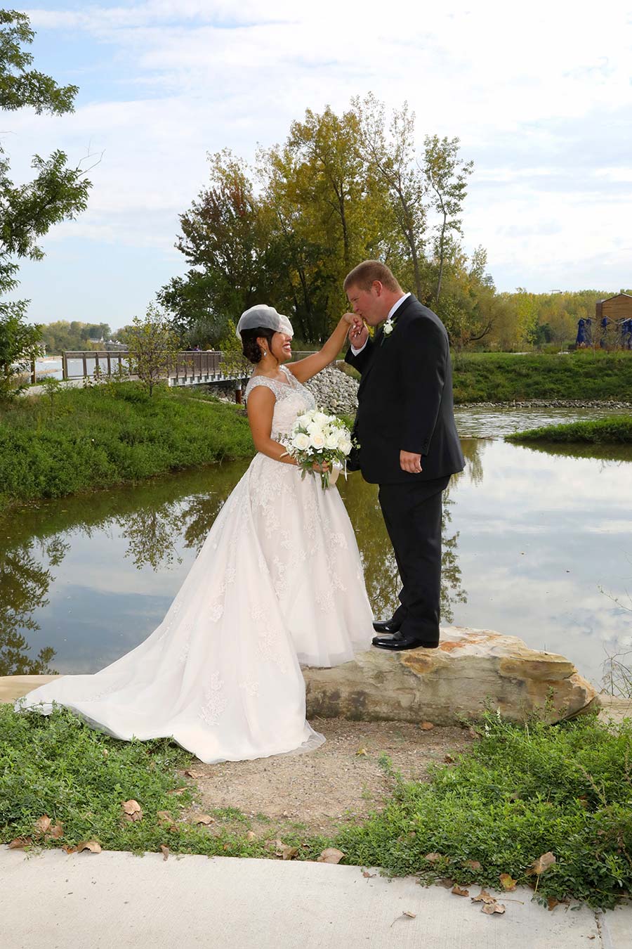 the groom give the bride a kiss on the hand while the couple stands on a large rock near the shores of the Maumee River