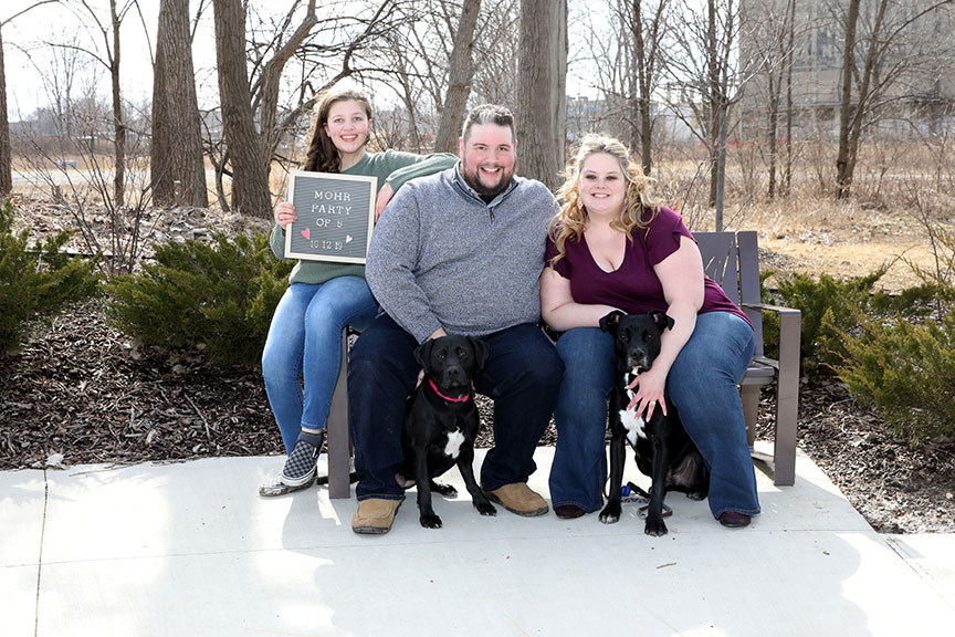 groom to be and bride to be with the groom's daughter and their two dogs sitting on a bench at Middlegrounds Park in downtown Toledo