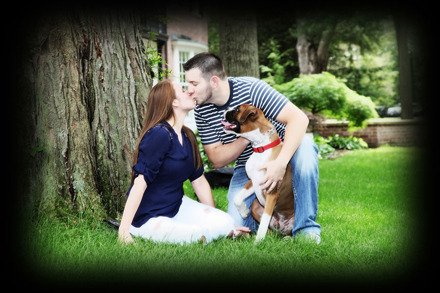 engaged couple kissing while sitting under a tree in the park with their dog