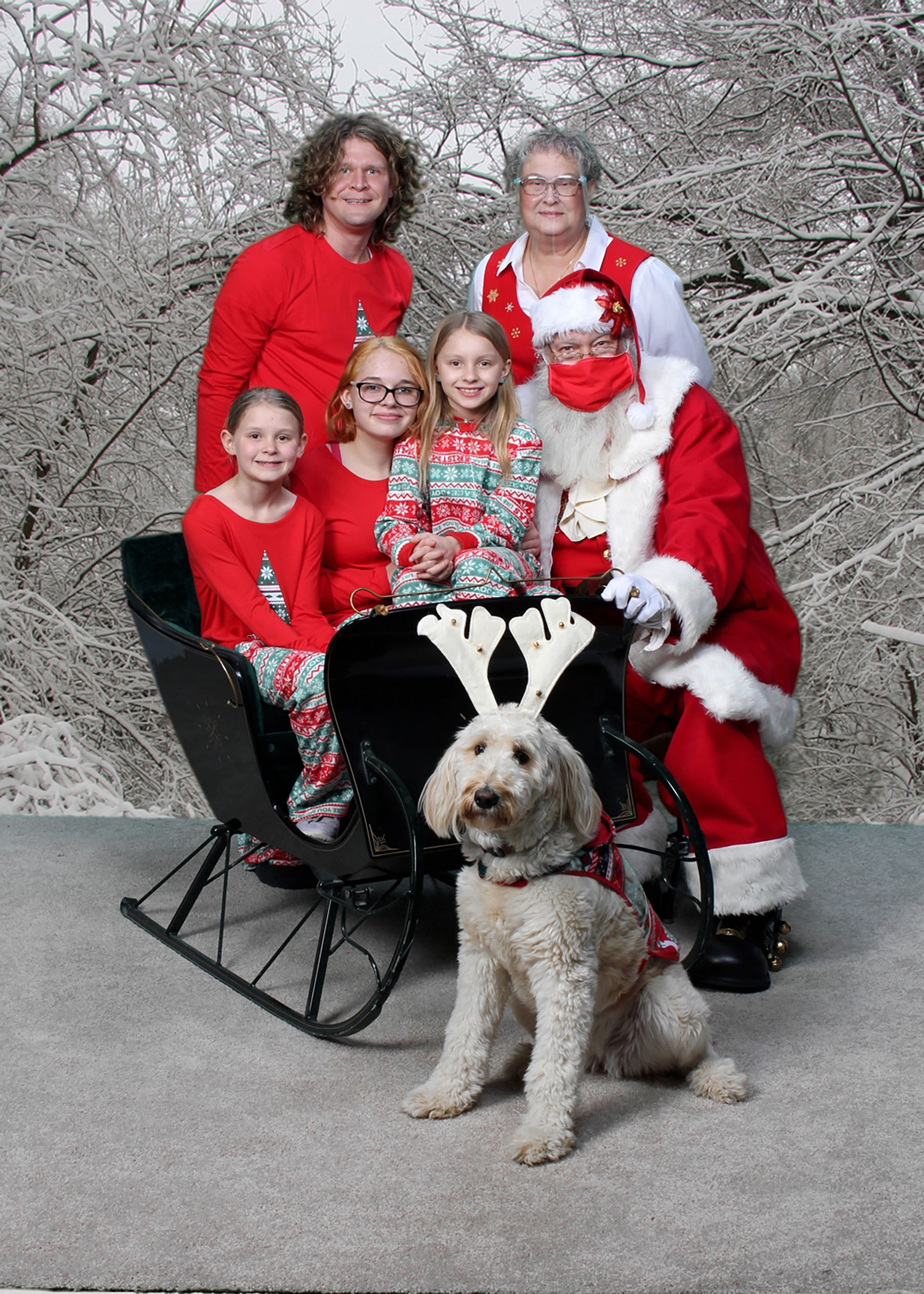 three sisters sitting in Santa's sleigh with their dad, Mrs. Claus and Santa behind them and the family dog dressed up like a reindeer
