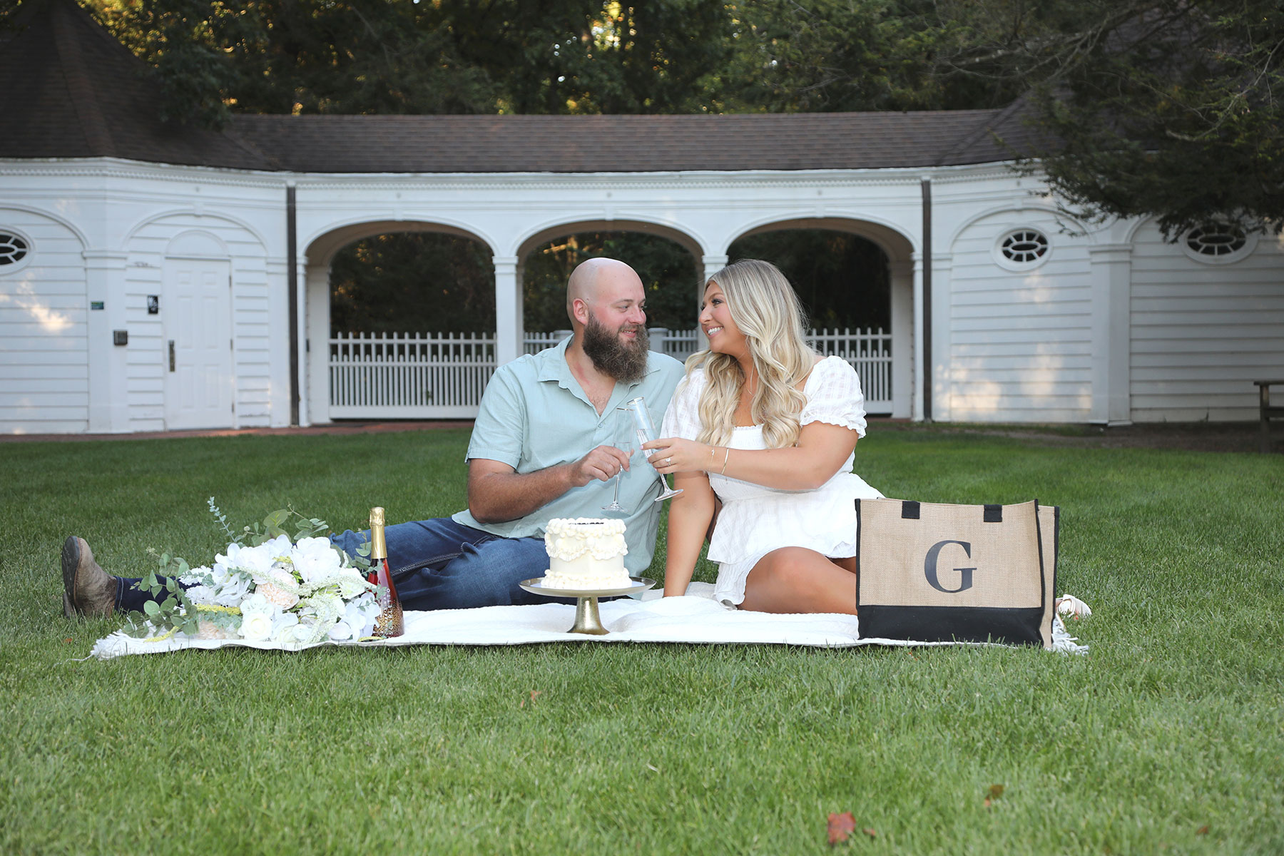 Engaged couple sharing a bottle of champagne and a small wedding cake picnic style on the lawn at Wildwood Metropark as part of the engagement photo session by Kurt Nielsen Photography